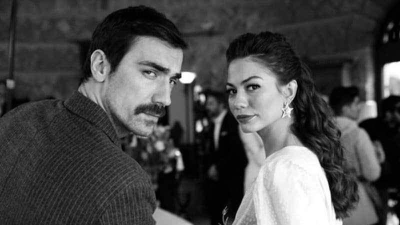 Ibrahim Çelikkol leaving the series dressed in black suit with moustache together with Demet Ozdemir wearing a white dress, both are looking over shoulder