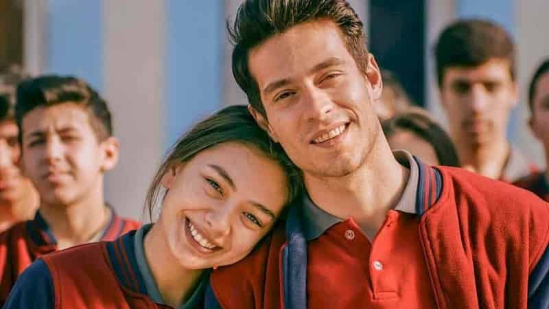 senden bana kalan, neslihan atagul wearing a red school uniform, smiling and laying her head on Ekin Koc, who is dressed with a red jacked and laying his head on her head.