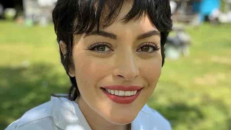face close up of ezgi mola smiling, with short dark hair and red lips, wearing a white t-shirt ayten