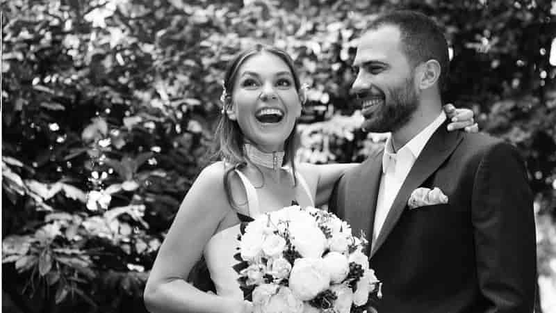 on left asli enver smiling, wearing a white wedding dress with a bouquet of white roses hugging Berkin Gökbudak who wears a black suit