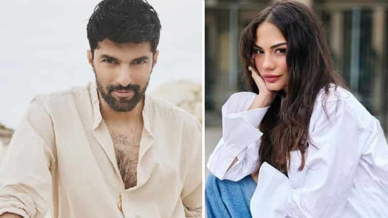 on left, Engin Akyürek wearing a beige shirt with dark hair and bear looking foward, on right Demet Özdemir wearing blue jeans with a white large blouse with curly long black hair, holdin her left hand under chin new series benim adim farah