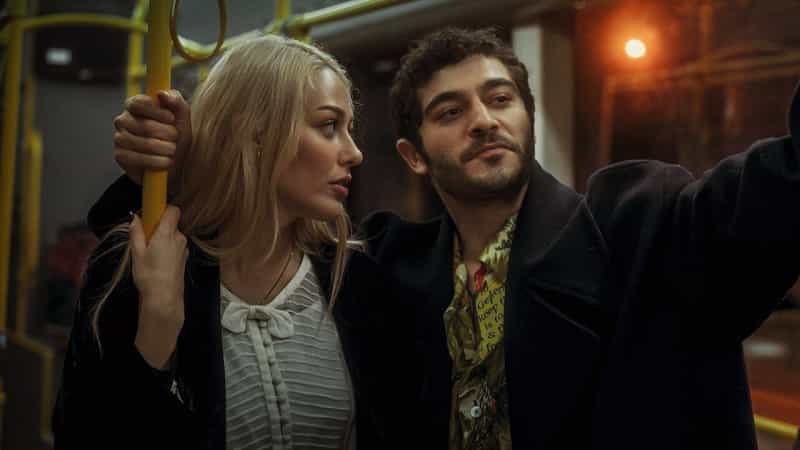 kal dont leave, in a train Dilan Çiçek Deniz wearing a white and black line shirt with black jacket and blonde hair looking on her right at Burak Deniz smiling wearing a yellow t-shirt and black jacket