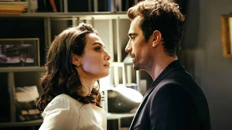 Season 2 Kuş Uçuşu As the Crow Flies, Birce Akalay with dark brown hair wearing a white blouse standing in front and looking into the eyes of İbrahim Çelikkol who wears a blue suit
