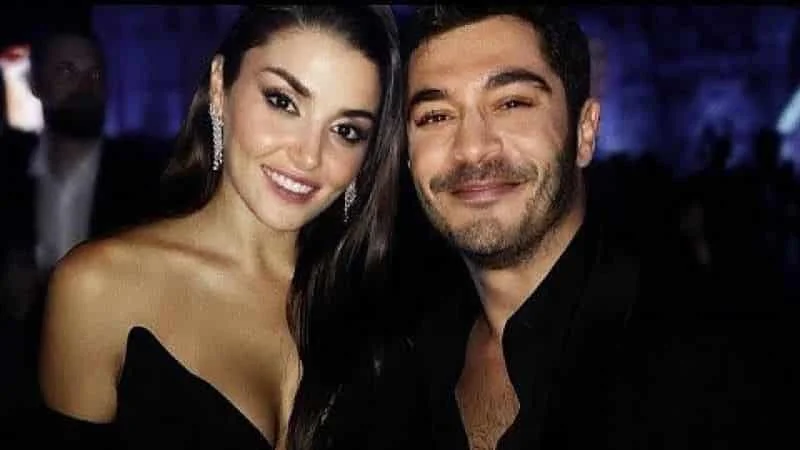 Hande Erçel with long black hair wearing a black dress with a rich decolletage next to Burak Deniz who smiles and wears a black shirt with her chest out
