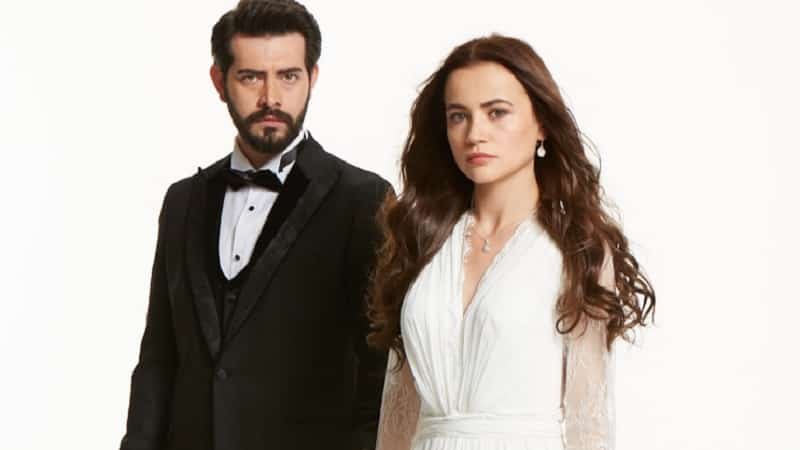 Kan Çiçekleri series youtube dizi turkish, white background with Yağmur Yüksel on the right side wearing a white dress and long brow hair, next to her is Barış Baktaş wearing a black costume with a bow