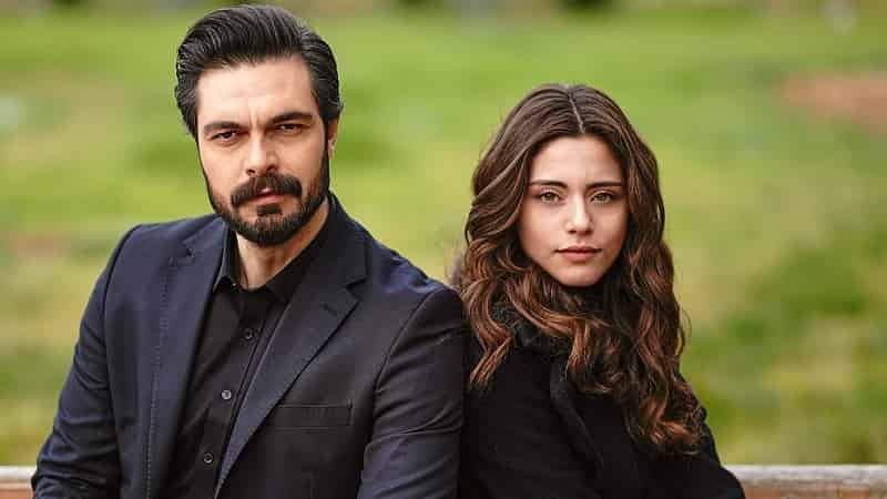 Halil Ibrahim Ceyhan yaman wearing a blue navy suit with black ahair and beard next to Sıla Türkoğlu wearing a black sweater with curly brown hair on a green background, emanet dizi series