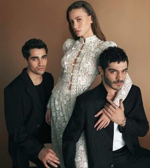 leading actors of Shahmaran series on netflix, Burak Deniz wearing black suit in front of Serenay Sarikaya who wears white dress, and keeps her hand on him with Mert Ramazan Demir wearing black suit behind her