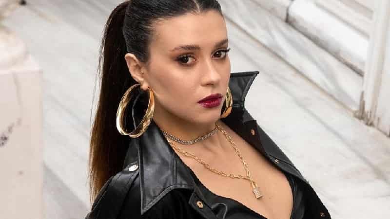 cem Tatlıtuğ stands up for Buçe Buse Kahraman with red lips, wearing a leather jacket with big gold and round earings and her hair in a pony tail