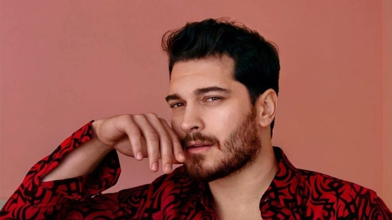 Çağatay Ulusoy wearing a red shirt holding his right hand to the chin, with bear and short hair preparing for new turkish tv series gaddar by ay yapim