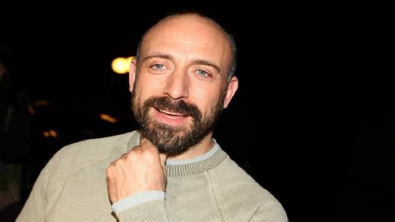 Halit Ergenç bald and beard wearing a grey shirt, with his hand under chin in the dark Stars in New Turkish Series ‘Adsız Aşıklar’ on Netflix